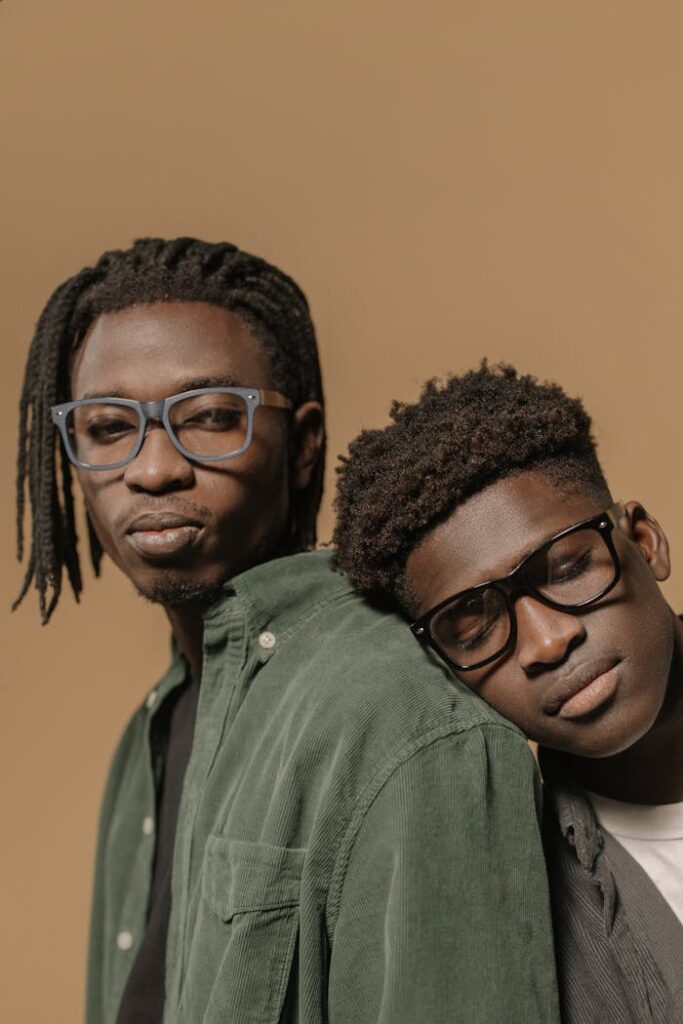 Close-up portrait of two stylish men wearing eyeglasses, one resting his head on the other's shoulder, against a beige background.